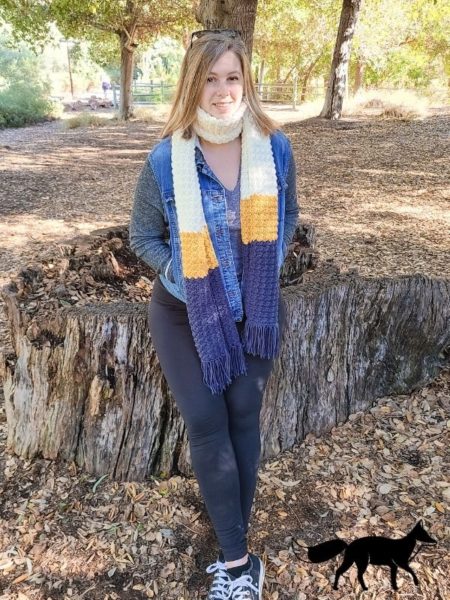 Image shows a woman sitting on a large tree stump in the wilderness. She is wearing the easy crochet scarf, showing how long and wide it is.