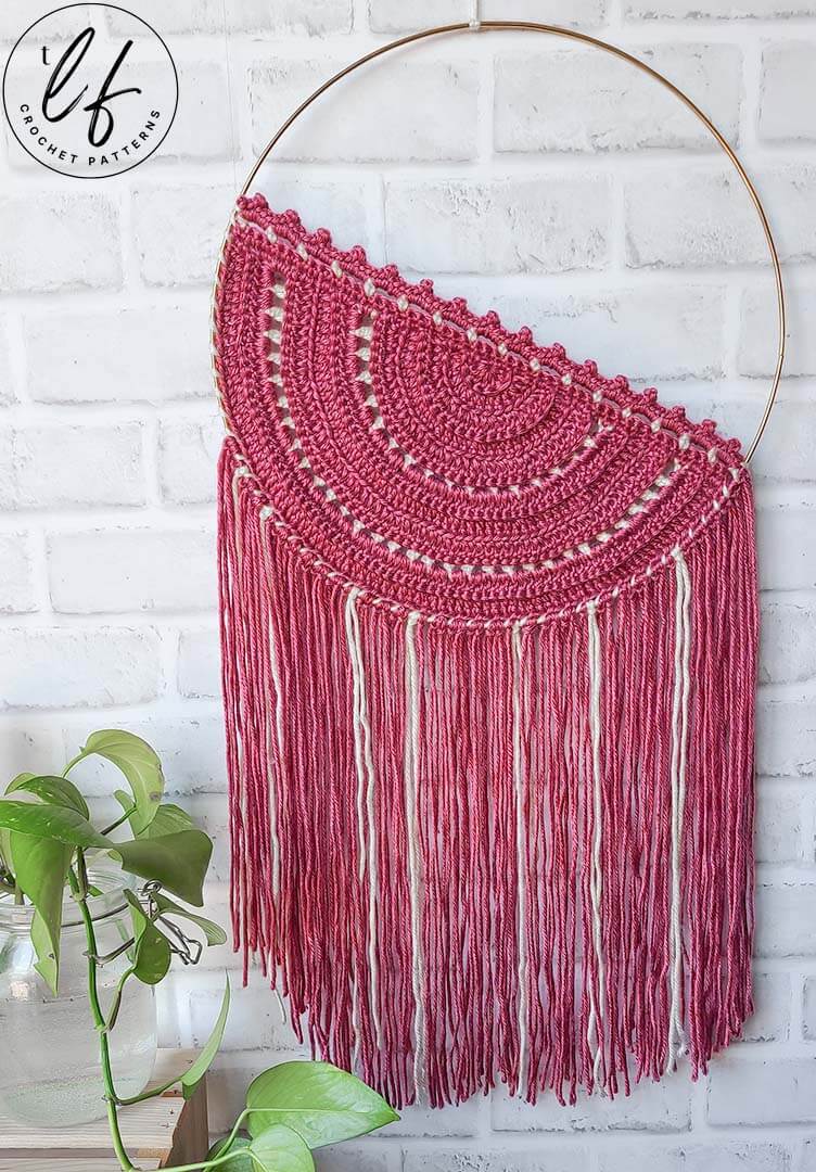 This image shows the crochet wall hanging on a white background, with a plant sitting in the lower left hand corner.