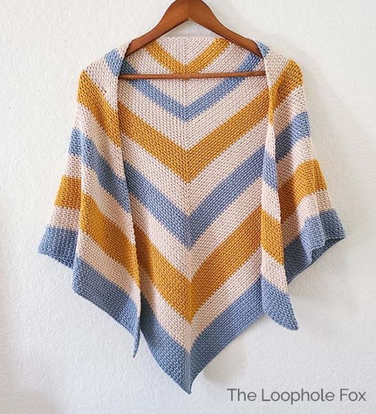 This image shows the moss stitch crochet shawl hanging on a wooden hanger on a white wall.