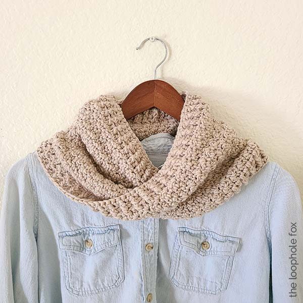The crochet infinity cowl pattern is shown finished, hanging on a wooden hanger with a light blue denim over shirt.
