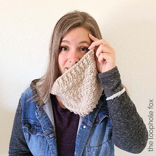 The crochet infinity cowl pattern is shown finished, worn by a woman. Woman is holding the edge of the cowl up to show the width. A fun, silly picture.