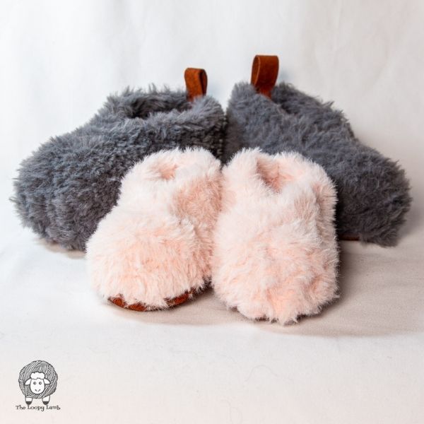 This image shows the Rustic Fur Crochet Slippers by The Loopy Lamb. These slippers are shown in 2 sizes, one large and one small. The small size is in a pink color and facing forward, the large size is behind facing slightly outward and made in grey. A white background finishes the image for these cute crochet projects. 