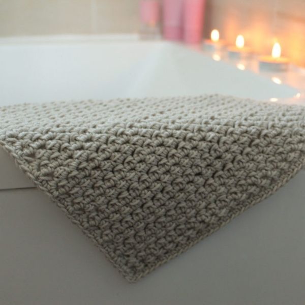 This image shows the Blossom Washcloth by Blue Star Crochet. The washcloth is shown laying on the side of a bathtub with candles in the background. 
