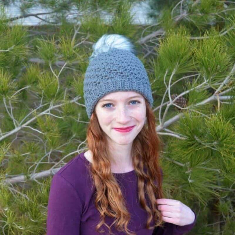 This image shows the Twilight Beanie worn by a woman who is standing in front of tree branches. The beanie is made in a blueish gray color with a white pom pom on top.