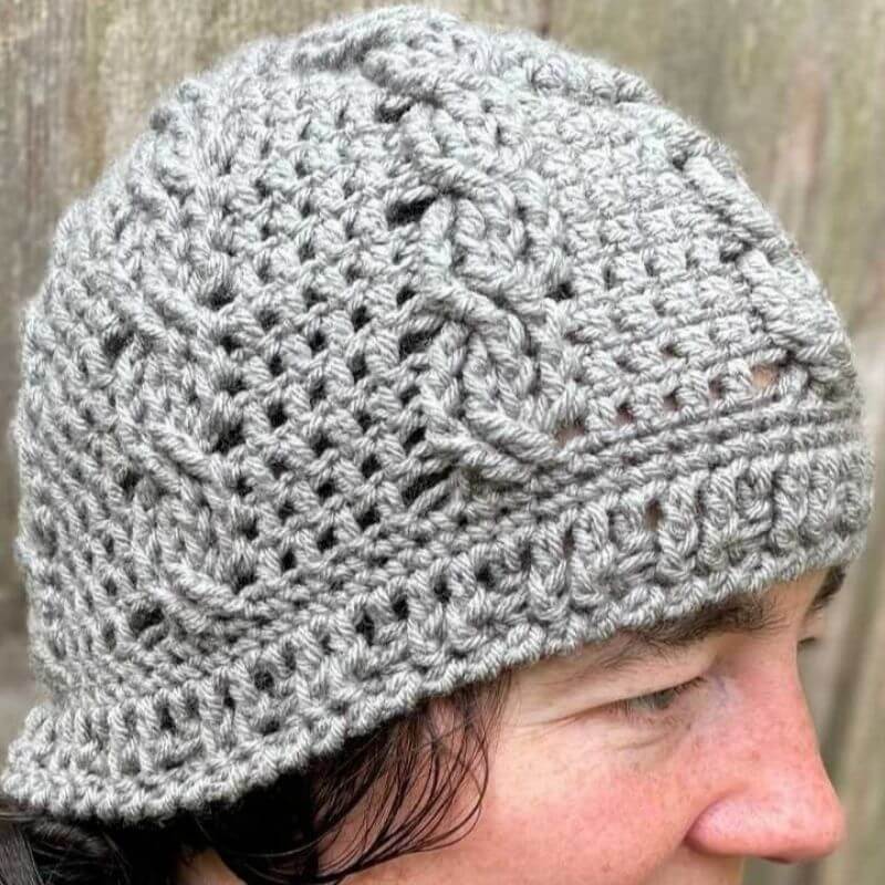 This image shows the Cozy Cabled Beanie worn by a woman who is standing in front of a wooden fence. The beanie is made in a grey color and shows the cabling. 