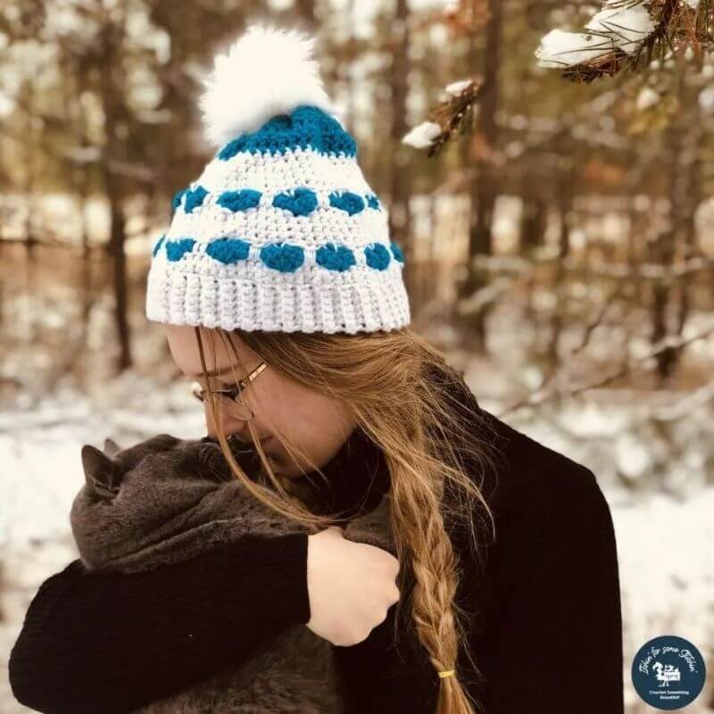 This image shows the You'll Always Be in My Heart beanie, one of the free crochet hat patterns in this round up, worn by a woman who is outside in a snowy backdrop. The beanie is made in white with blue hearts.