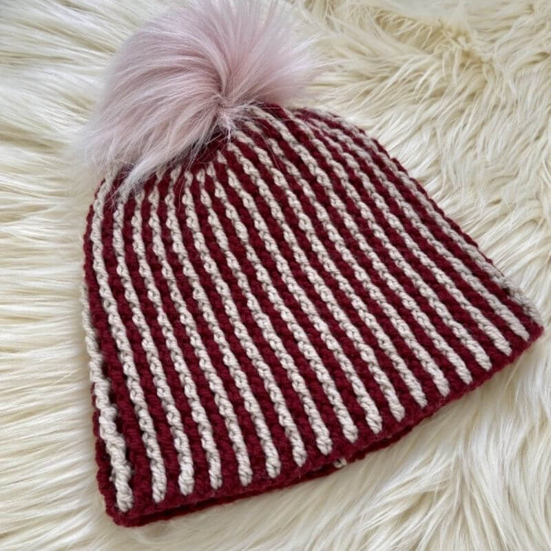 This image shows the Anton Beanie, one of the free crochet hat patterns in this round up, laying flat on a fluffy white surface. The beanie is vertically striped in red and grey.