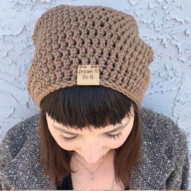 This image shows the Dreamer Beanie, one of the free crochet hat patterns in this round up, worn by a woman outside standing in front of a grey wall. She is looking down to show more of the beanie. The beanie is made in a light brown color and does not have a brim.
