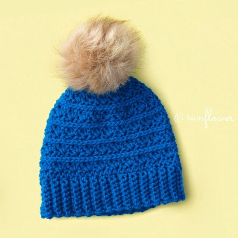 This image shows the Samantha's Hope Beanie laid flat on a yellow background. The beanie is made in blue and has a pom pom on top.