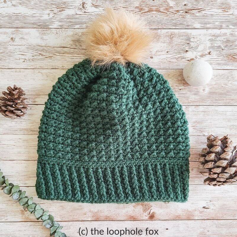 This image shows the Arctic Beanie laying flat on wood background. It is in a dark green color with a tan colored pom pom on top. Around the beanie are pinecones.