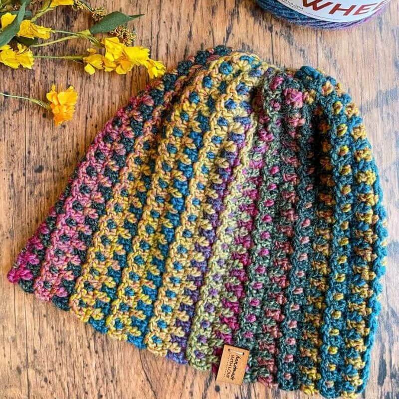 This image shows the Mountain Trail Beanie laid flat on a wooden background with yellow flowers peaking in at the top left corner. The beanie is made in a variegated yarn that has purple, yellow, blue, maroon and green. 