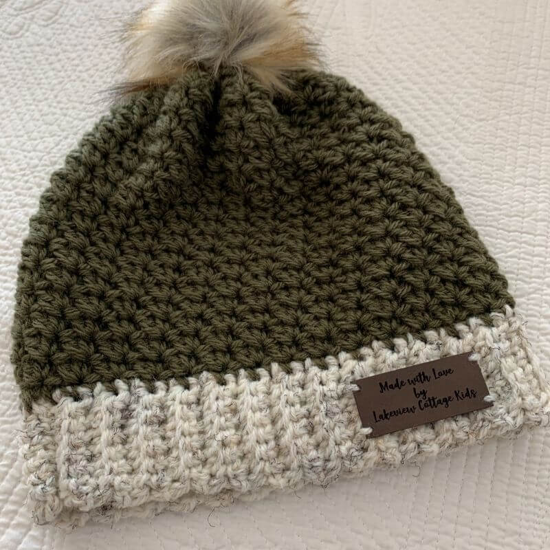 This image shows the Up North Beanie laid flat on a white, textured background. The beanie is made in an olive green and the brim is made in a white tweed.