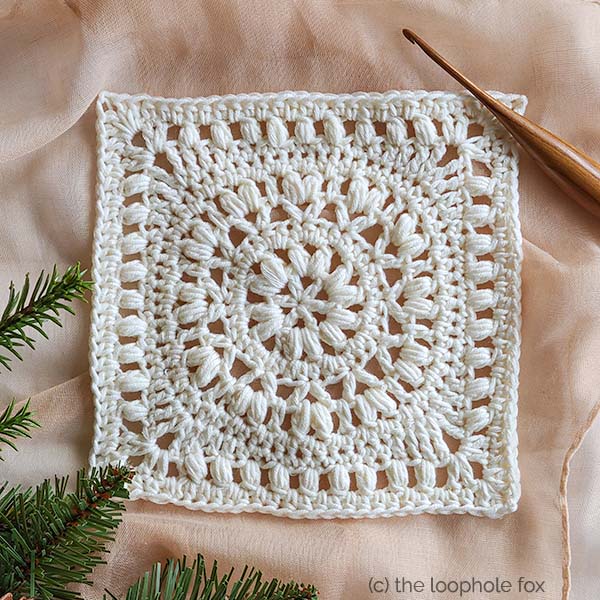 This image shows the Marguerite Square, a square crochet square pattern, worked twice. Once in white yarn and one in olive green yarn. The olive square sits behind the white square, which is situated diagonally. A small amount of foliage peaks up from the bottom left corner and a wooden crochet hook sits diagonally in the top right corner.