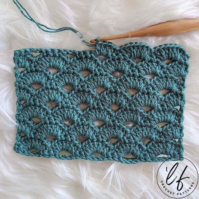 This image shows a swatch of the Crochet Arcade Stitch. The swatch is laying flat on a fluffy white background and is made in a sage green color. The swatch has a row half worked, with the wooden crochet hook still in the stitch.
