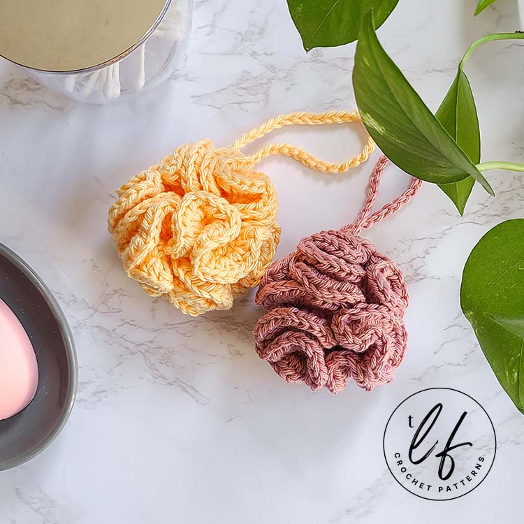 This image shows the crochet bath pouf pattern, worked twice - once in yellow and once in pink - with a bit of pothos plant and other bathroom necessities around for a staged picture.