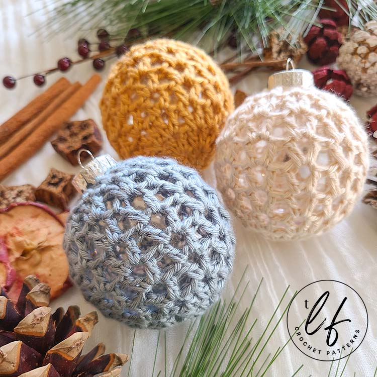 3 crochet Christmas baubles (one white, one blue, one yellow) shown laying flat on a white background. Picture is taken from an angle from the bottom left side, to show depth. An assortment of Christmas related items such as pine cones and cinnamon sticks decorates the photo.