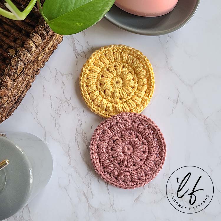 This image shows two samples of the crochet face scrubby pattern. One is made in yellow and the other in pink. They are laying on a marble background with typical bathroom accessories around them, like a basket, a bar of soap in a dish, etc.