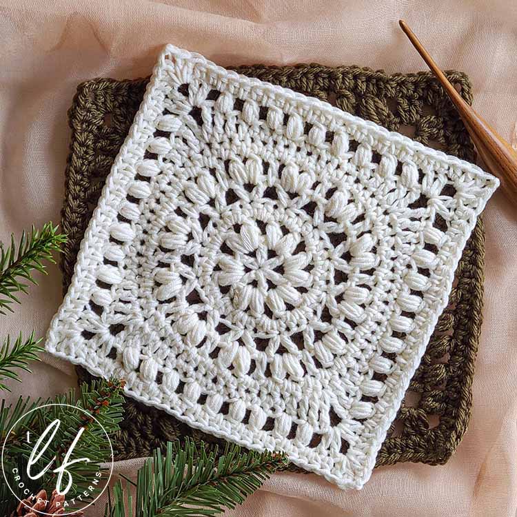This image shows the Marguerite Square, a white square crochet pattern, laid flat on a textured background of taupe fabric. A bit of foliage peaks out from the bottom left corner and a wooden crochet hook sits diagonally in the top right hand corner.
