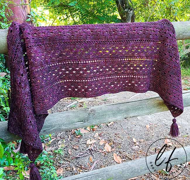 This image shows the crochet rectangle shawl pattern sample laying draped on a wooden fence with trees and bushes in the background.