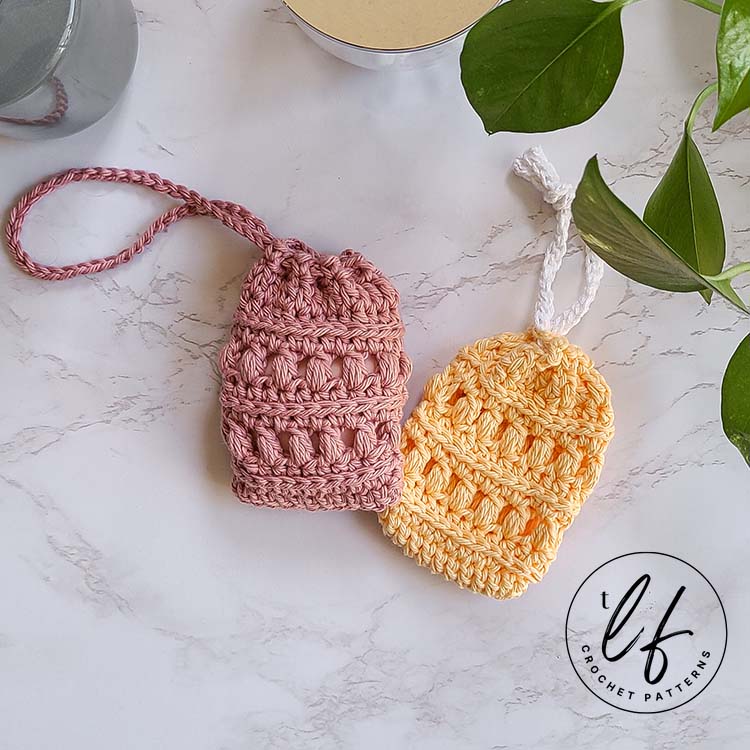 This image shows the crochet soap saver pattern made twice - once in pink and once in yellow. They're laid next to each other on a marble background, with green plant vines in the upper right hand corner and bathroom essentials peaking in at the top.