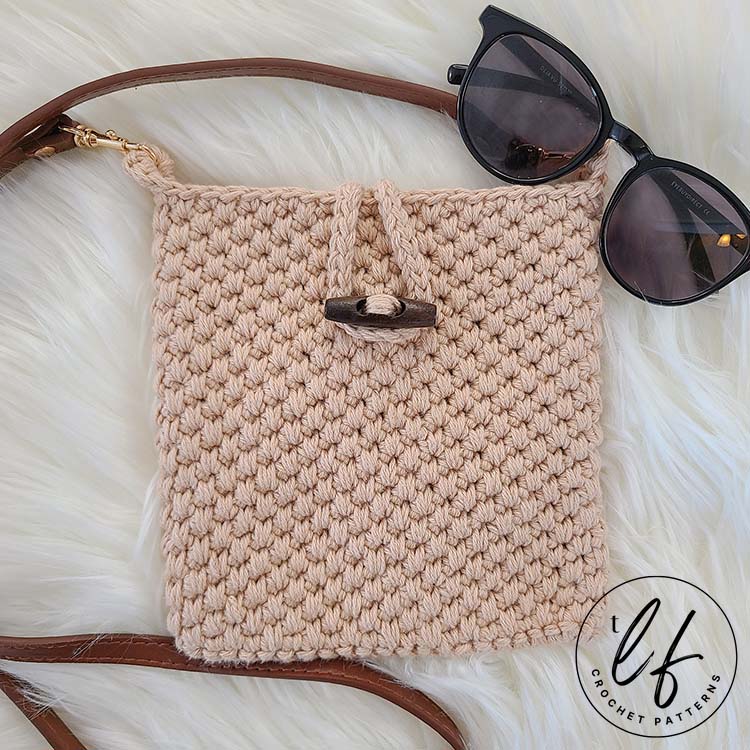 Image shows the crochet crossbody bag pattern sample laid flat on a fluffy white surface. The loop is closed around a toggle button to show the closure. A pair of sunglasses sit in the upper right hand corner of the bag.