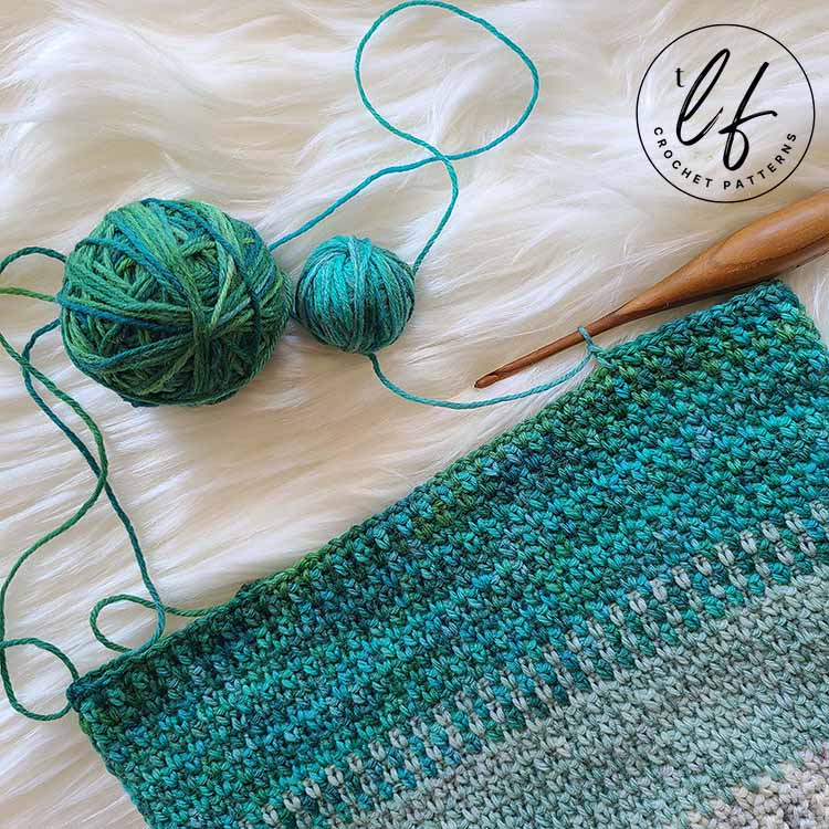 This is a work in progress image. This image has the beanie half way completed, laying flat. A crochet hook is still in one of the stitches, and two balls of colors are sitting above the work.