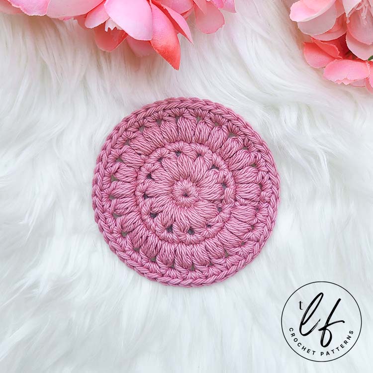 This image shows the crochet face scrubby pattern sample laying flat on a fluffy white background. The face scrubby is made in pink and a small amount of flower petals line the top of the picture.