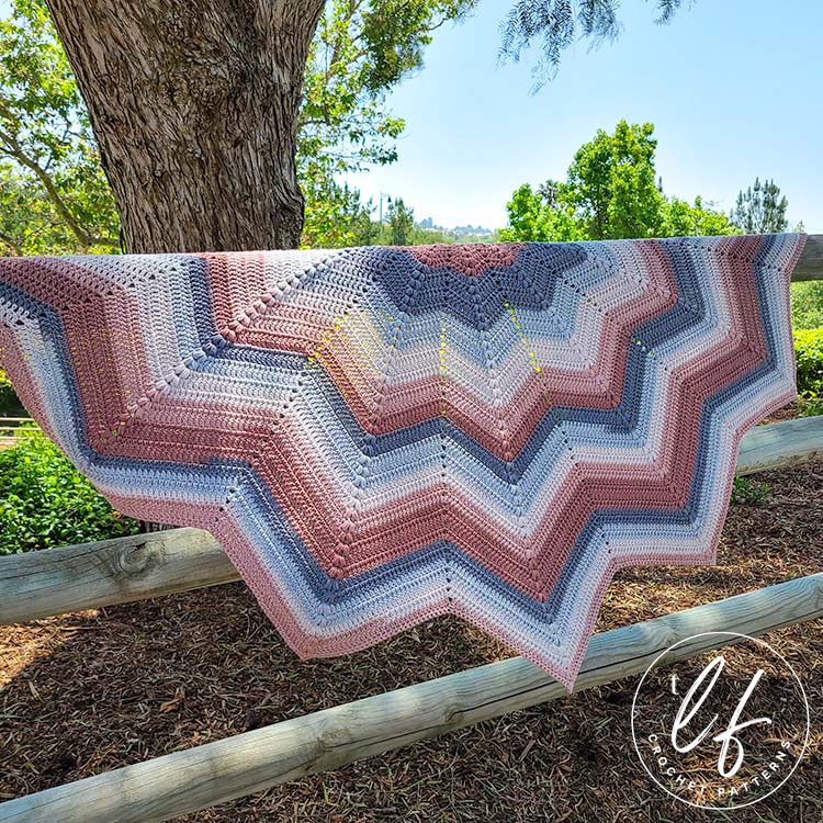 This image shows the crochet chevron shawl hung on a wooden fence, with trees and other foliage in the background. The shawl is spread wide so you can see each peak and valley of the chevron. Sun hits the top of the shawl, showing off the puff stitches.