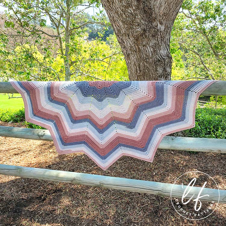 This image shows the crochet chevron shawl hung on a wooden fence, with trees and other foliage in the background. The shawl is spread wide so you can see each peak and valley of the chevron.