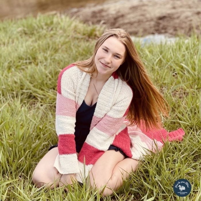 This image shows the Lyndie Cardi, one of the crochet projects for summer, worn by a young woman who is sitting in the grass.