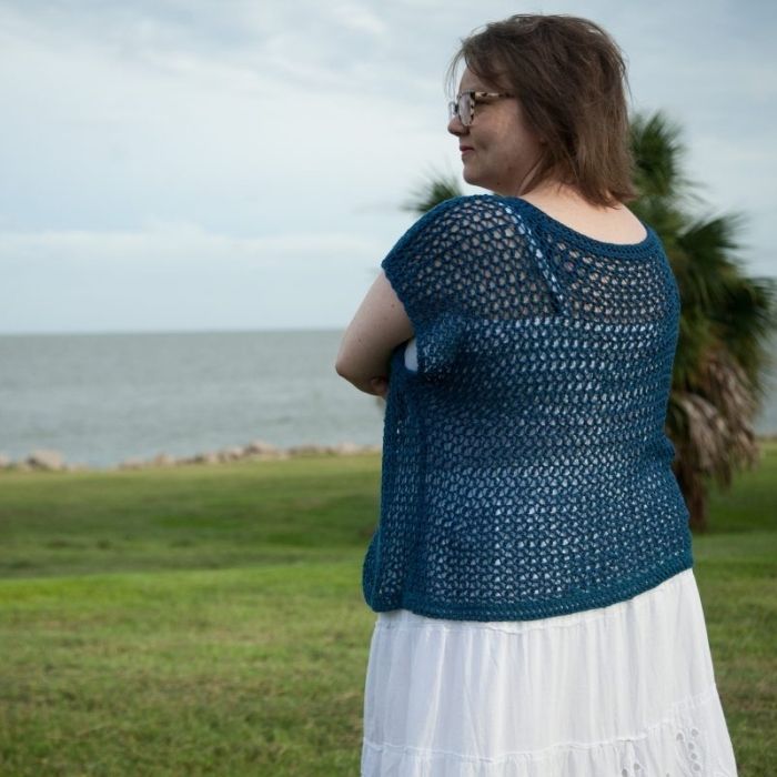 This image shows the Shirl Top, one of the crochet projects for summer, worn by a woman who is standing in a field in front of a body of water.