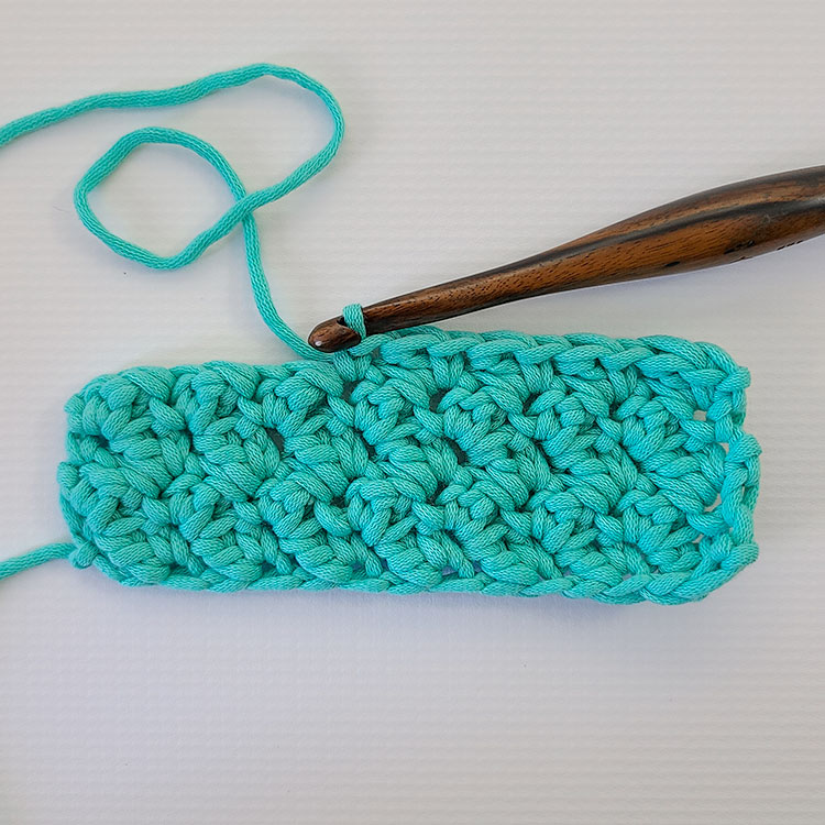 This image shows a swatch of the crochet suzette stitch. The last row is worked halfway with the hook still in the yarn. The yarn is a bulky weight in a teal color and the hook is dark wood. It is on a white background.
