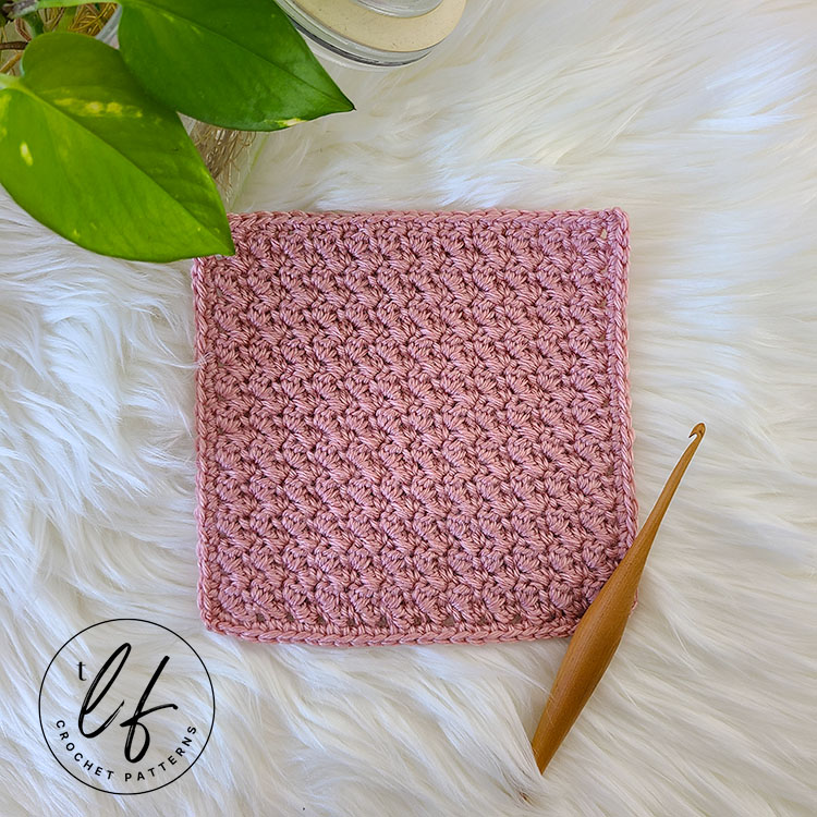 This image shows a 7 inch by 7 inch square crocheted using the crochet Suzette Stitch. The square is in a soft pink color and is laying on a fluffy white background. A crochet hook sits in the bottom right corner and a bit of a plant peaks in from the top left corner.