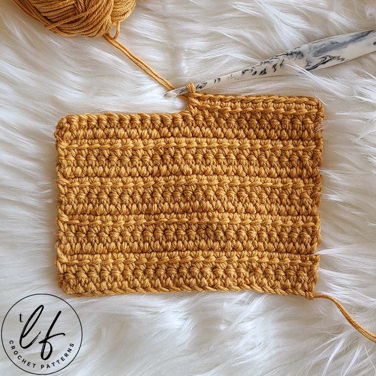 This image shows a swatch of the Linked Double Crochet laid flat on a fluffy white background. The swatch is crocheted in a golden yarn and the last row is half worked, with the hook still in the working loop.