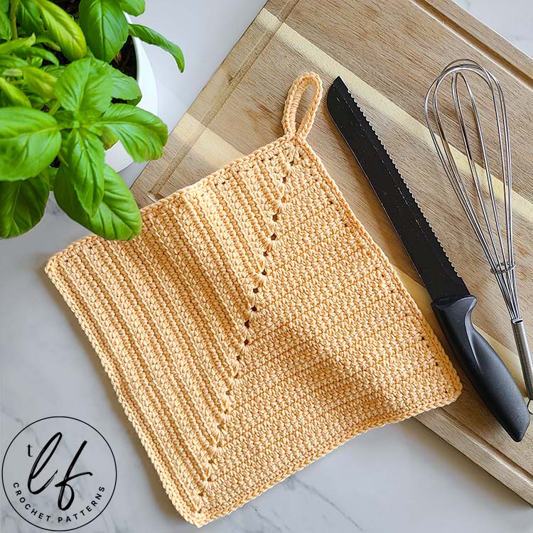This image looks down at a marble counter top. The crochet kitchen towel is laying half on and half off a wooden cutting board. A bread knife and whisk are also on the cutting board. A basil plant peaks in at the top left.