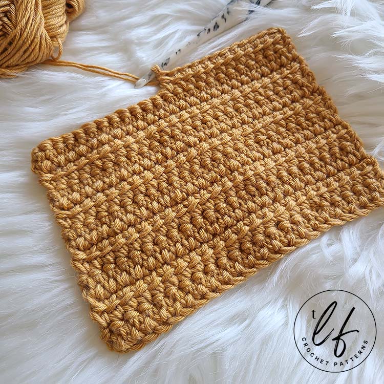This image shows a swatch of the Linked Double Crochet laid flat on a fluffy white background. The swatch is crocheted in a golden yarn and the last row is half worked, with the hook still in the working loop. The swatch is laying at an angle.