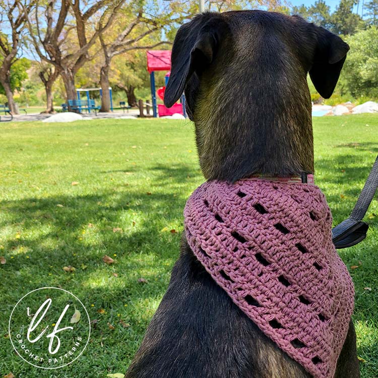 This image shows a medium sized black dog wearing the lumina bandana. The image is taken from behind, so you can see the dog sitting and the bandana on their neck. The dog is sitting on a shaded grass field and she is looking out at a playground and trees.
