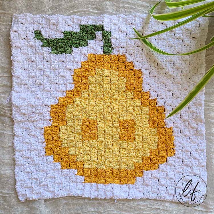 This image shows pear the corner to corner square laid flat on a white background. The pear is crocheted in yellows with a green stem. A little bit of a spider plant peaks in on the top right corner.