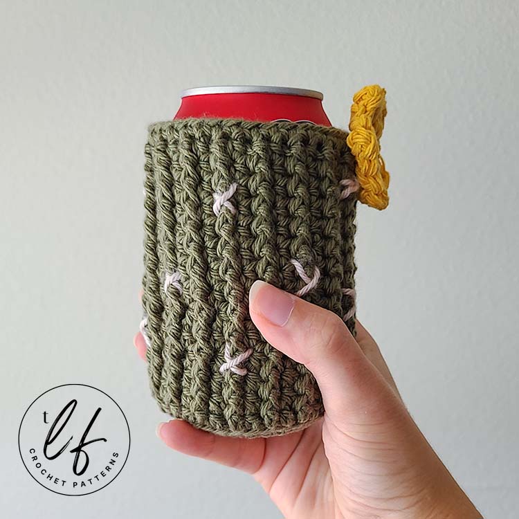 This image shows a hand holding the crochet can cozy in front of a white wall. The can cozy is on a can so it is displayed as it would be used. It is styled after a cactus, so the cozy is ribbed with front post half double crochets and made in green. Little white X's create the spines and a yellow crochet flower is sewn to the top.