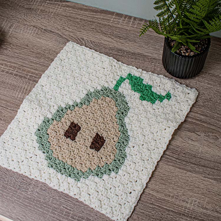 This image shows the pear corner to corner square as crocheted by Helen of Sunflower Cottage Crochet. The square is laid flat on a wooden desk and the picture is taken at a diagonal. The pear is crocheted in green and tan.