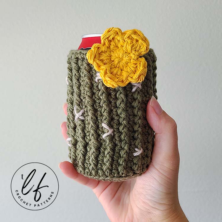 This image shows a hand holding the crochet can cozy in front of a white wall. The can cozy is on a can so it is displayed as it would be used. It is styled after a cactus, so the cozy is ribbed with front post half double crochets and made in green. Little white X's create the spines and a yellow crochet flower is sewn to the top.