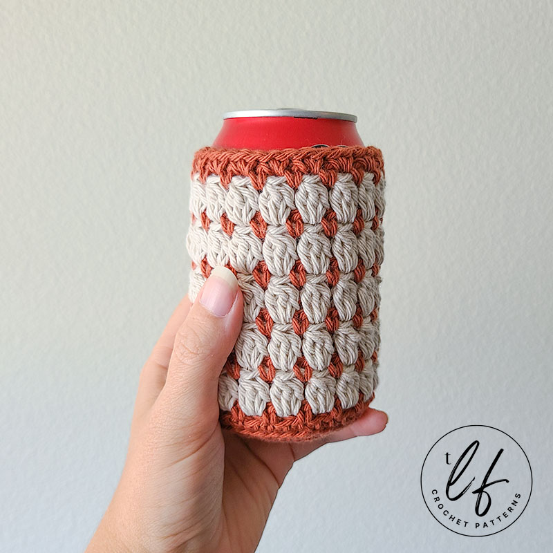 This image shows the crochet can cozy free pattern sample being held in front of a white wall. The can cozy is on a standard soda can and is made in both a cream color and a burnt orange color.