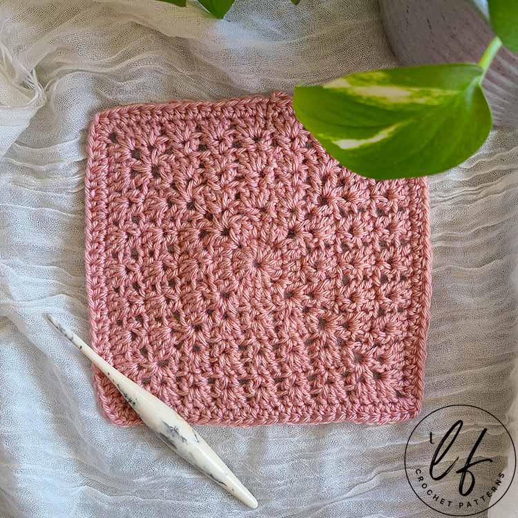 This image shows the modern crochet granny square sample laid flat on a white fabric background. The square is made in a soft pink color and a bit of a plant peaks in on the top right corner.