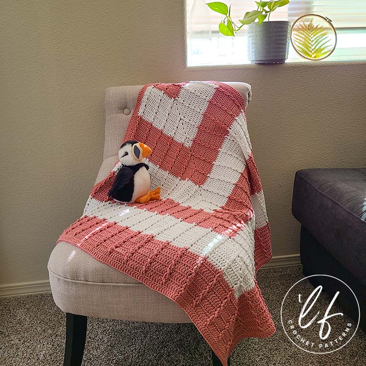 This image shows the modern crochet baby blanket laying draped on a white fabric chair with a stuffed puffin sitting on it. The chair is situated in front of a window, with a plant sitting in the sill. 