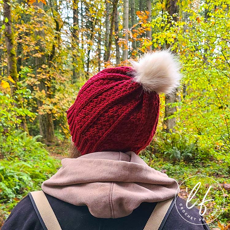 This image shows the free crochet beanie pattern sample being worn by a woman. The picture is taken from behind and the woman is looking slightly to the left. You can see the full beanie clearly. The background is trees and foliage. 