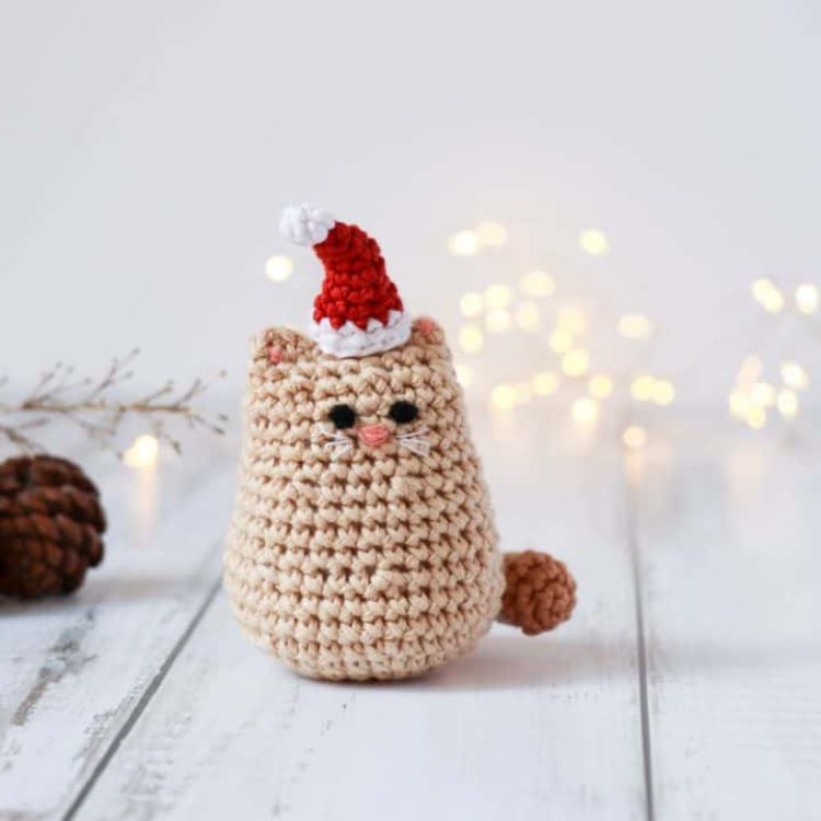 This image shows the next pattern, which is the Itty Bitty Christmas Kitty as described above. The image amigurumi cat sitting on a wooden surface with a white background. In the background there are fairly lights and pinecones.