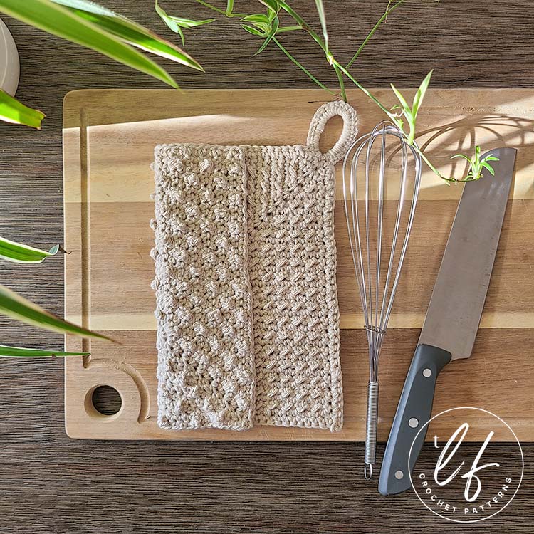 This image shows the crochet dishcloth pattern on a wooden cutting board with a whisk and knife next to it. The crochet wash cloth is folded so it is slightly overlapping itself, showing the different texture on either side.