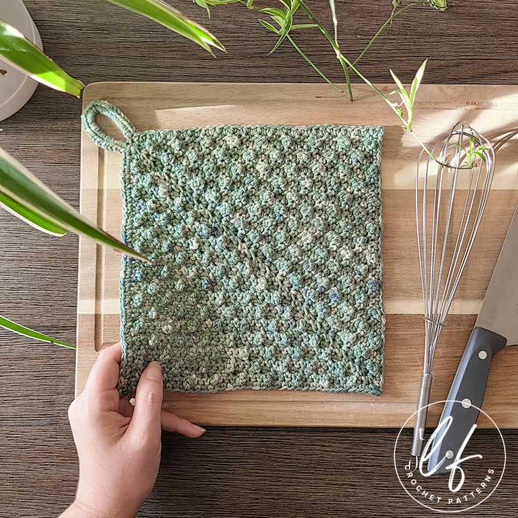 This image shows the crochet dishcloth pattern sample made in green. The dishcloth is laying on a wooden cutting board and a hand is holding the corner of the cloth up slightly.