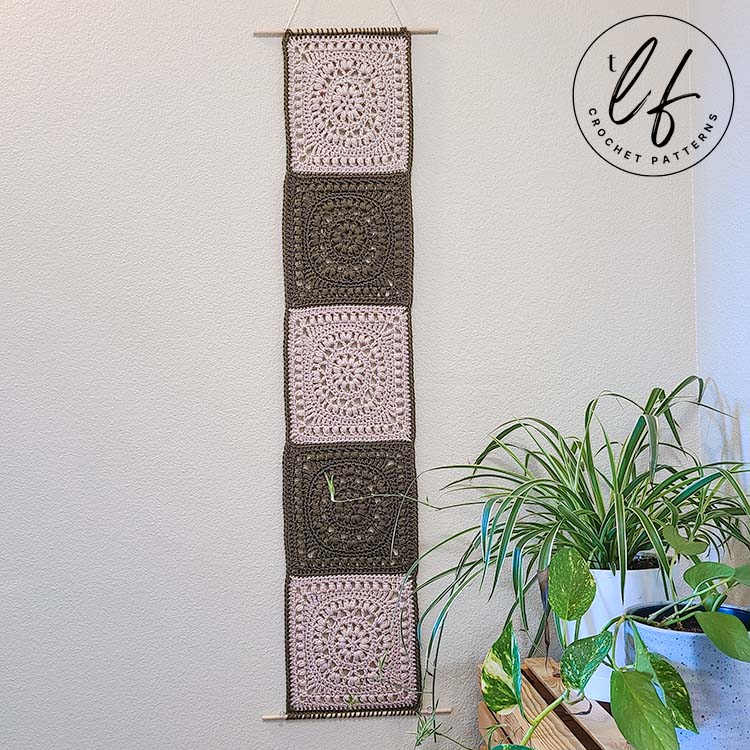 This image shows the granny square wall hanging completed using two colors, cream and olive. The finished project is hanging on a white wall next to plants.