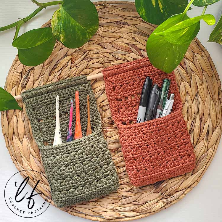 This image shows 2 finished baskets created from this Crochet Hanging Basket Pattern and are attached to one dowel side by side. The baskets are laying flat on a woven, natural placemat with a strand of pothos laying at the top. In the baskets are crochet hooks and pens.